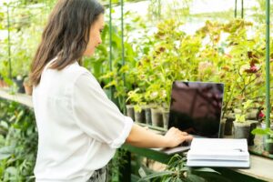 brunette woman on laptop in greenhouse managing her garden business